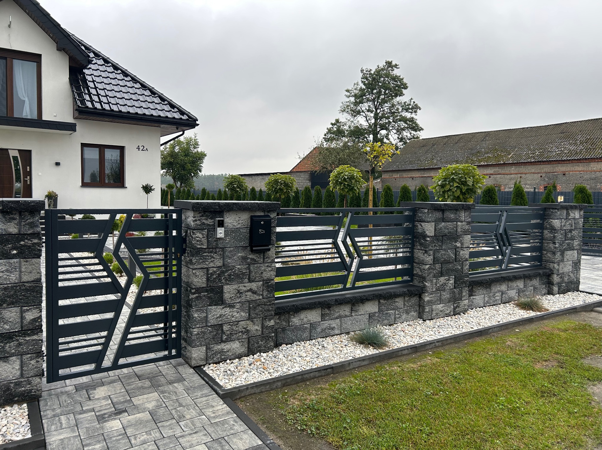Nowoczesne, grafitowe ogrodzenie panelowe z bramą wjazdową, z elementami dekoracyjnymi w kształcie rombów, osadzone na kamiennych słupkach, przed domem z czarną dachówką, w tle zabudowania...
