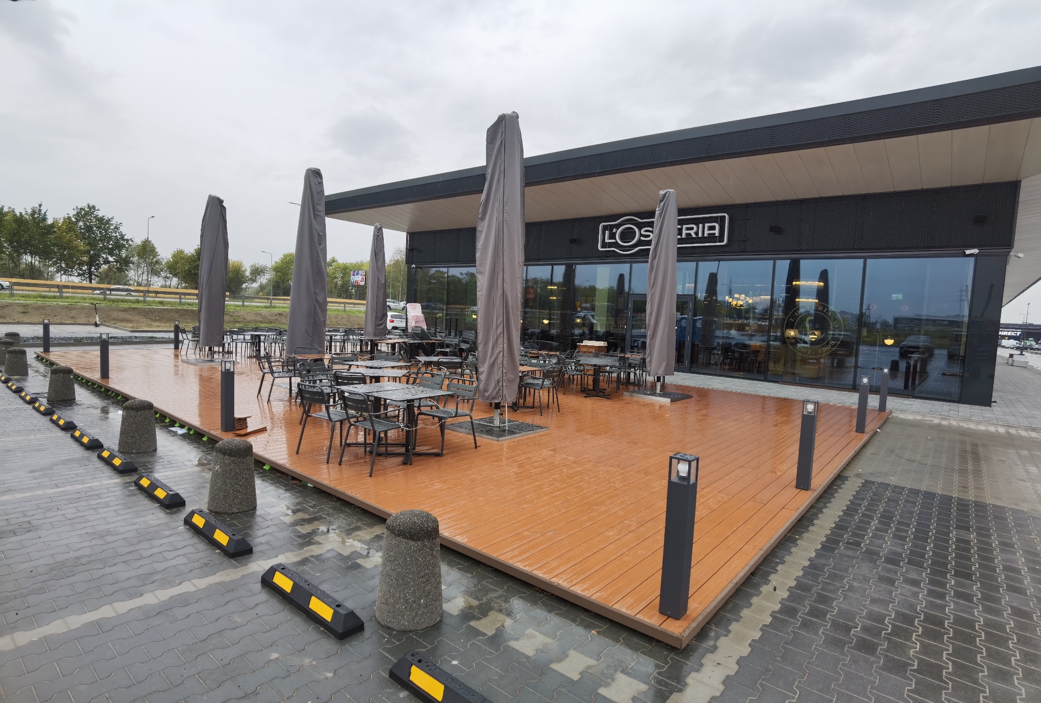 Zewnętrzny taras restauracji z drewnianą podłogą, szarymi stolikami i krzesłami, oraz parasolami w pokrowcach w pochmurny dzień. Widoczne betonowe słupki i odbojniki parkingowe.