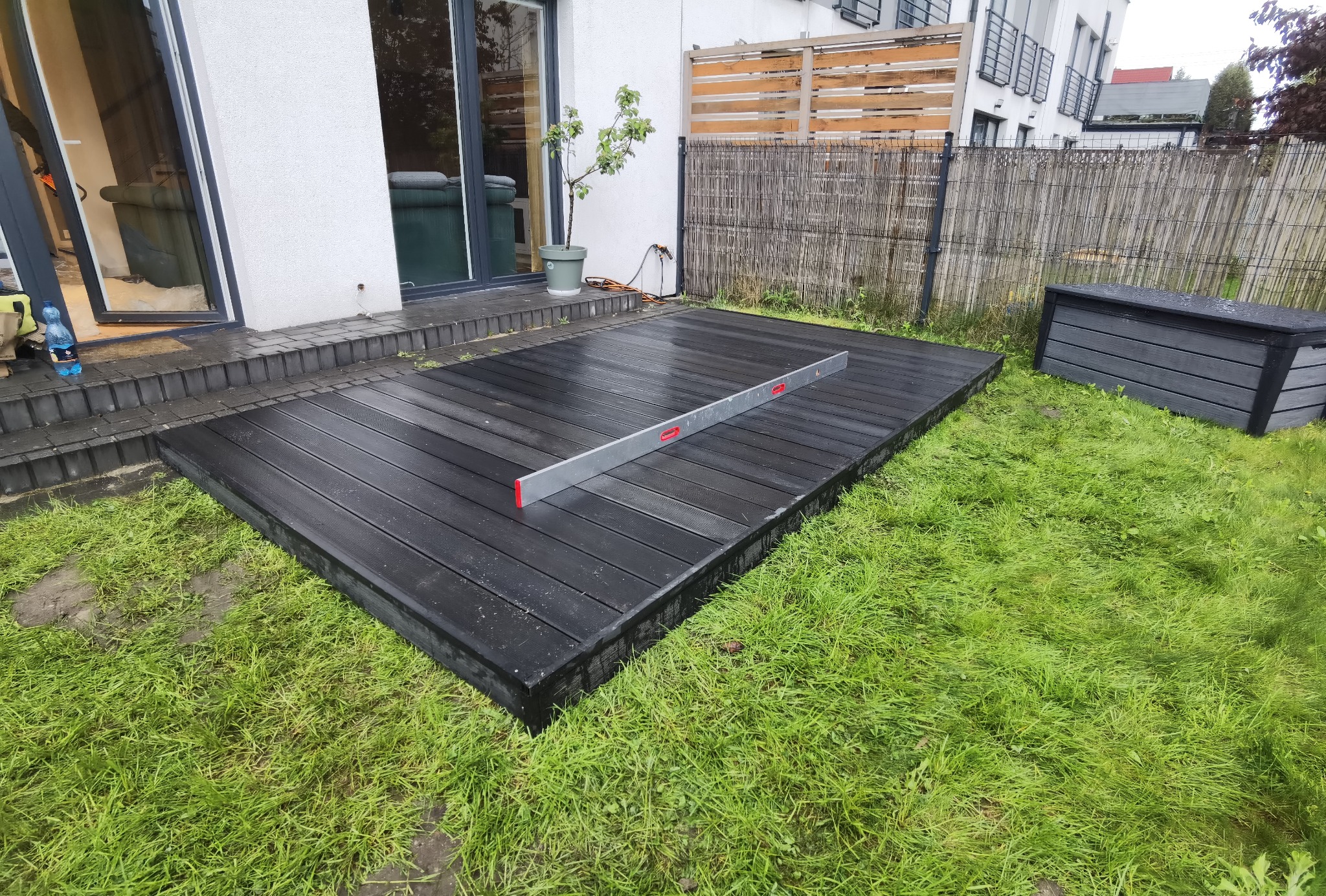 Ciemnoszary taras kompozytowy w trakcie montażu, poziomnica leży na deskach, widoczna skrzynia ogrodowa i trawnik.