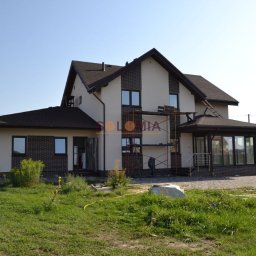 Solomia Sp. z o.o. - Dwukondygnacyjny dom z brązowym dachem i ceglanymi detalami, w trakcie wykończenia elewacji, z widocznym rusztowaniem i przygotowanym terenem wokół.