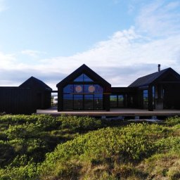 Solomia Sp. z o.o. - Czarny dom modułowy z dużymi oknami, widoczny z zewnątrz na tle zielonej roślinności i pochmurnego nieba, z logo firmy na szybie.