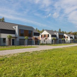 Solomia Sp. z o.o. - Szereg nowoczesnych domów z szarymi dachami i drewnianymi akcentami, ogrodzone siatką, z zadbanymi trawnikami i drzewami, widok z perspektywy ulicy w słoneczny dzień.