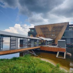 Solomia Sp. z o.o. - Nowoczesny budynek na wodzie z drewnianą fasadą i zadaszonym pomostem, widok z brzegu w pochmurny dzień.