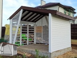 Biała, ażurowa konstrukcja z brązowym dachem w ogrodzie. Widoczne poziome listwy i fragment składowanego drewna wewnątrz. W lewym dolnym rogu dodano małą wizualizację 3D tej samej konstrukcji.