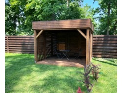 Drewniana konstrukcja altany z ażurowymi ścianami, wyposażona w stolik i dwa krzesła, ustawiona na trawniku przed drewnianym ogrodzeniem, otoczona zielenią drzew.