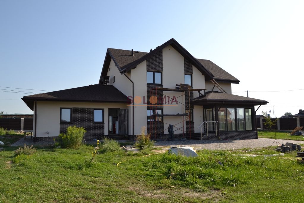 Dwukondygnacyjny dom z brązowym dachem i ceglanymi detalami, w trakcie wykończenia elewacji, z widocznym rusztowaniem i przygotowanym terenem wokół.