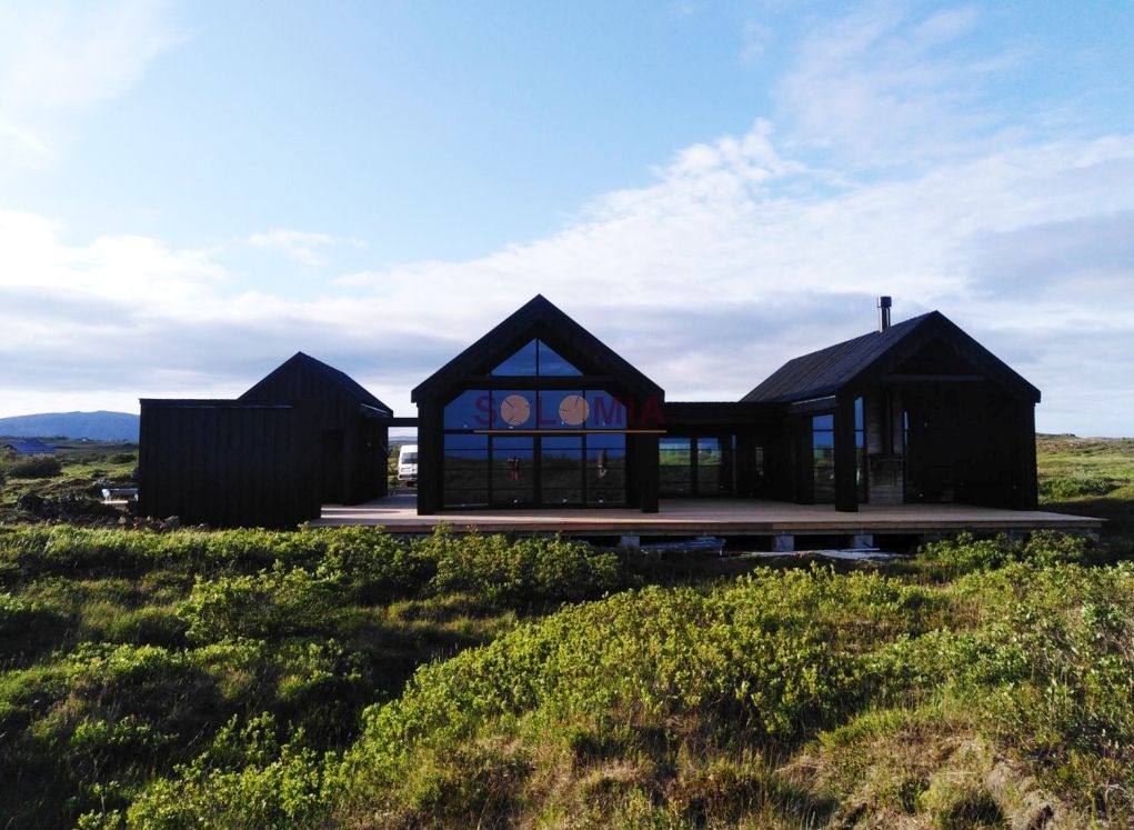 Czarny dom modułowy z dużymi oknami, widoczny z zewnątrz na tle zielonej roślinności i pochmurnego nieba, z logo firmy na szybie.