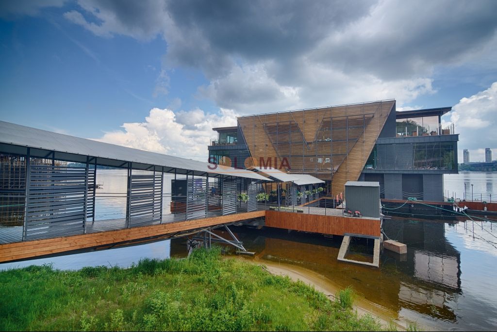 Nowoczesny budynek na wodzie z drewnianą fasadą i zadaszonym pomostem, widok z brzegu w pochmurny dzień.