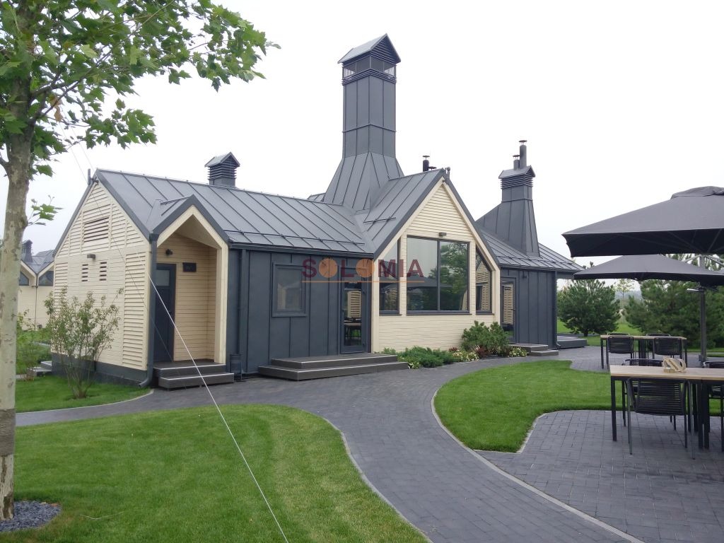 Nowoczesny budynek z elementami drewnianymi i metalowym dachem, otoczony trawnikiem i ścieżką z kostki brukowej. Widoczny fragment tarasu z parasolem i meblami ogrodowymi.