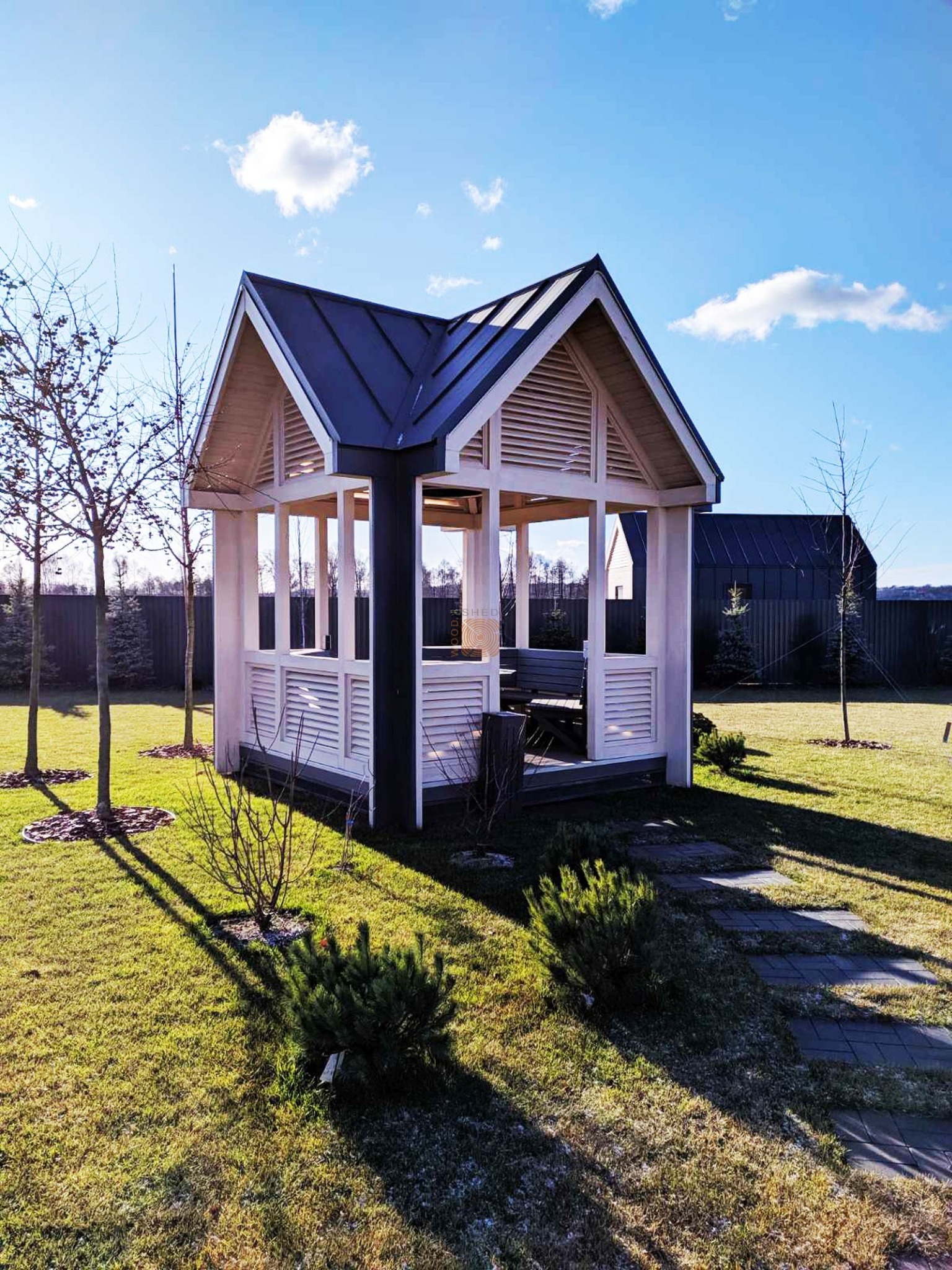 Biała altana z ciemnym dachem w ogrodzie, widok z zewnątrz w słoneczny dzień, z widoczną ścieżką z płyt i młodymi drzewkami.