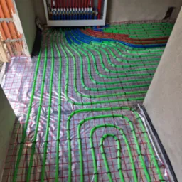 Rozłożone rury ogrzewania podłogowego w kolorze zielonym, niebieskim i czerwonym na folii z siatką, widoczna szafka rozdzielcza z zaworami i poziomica.