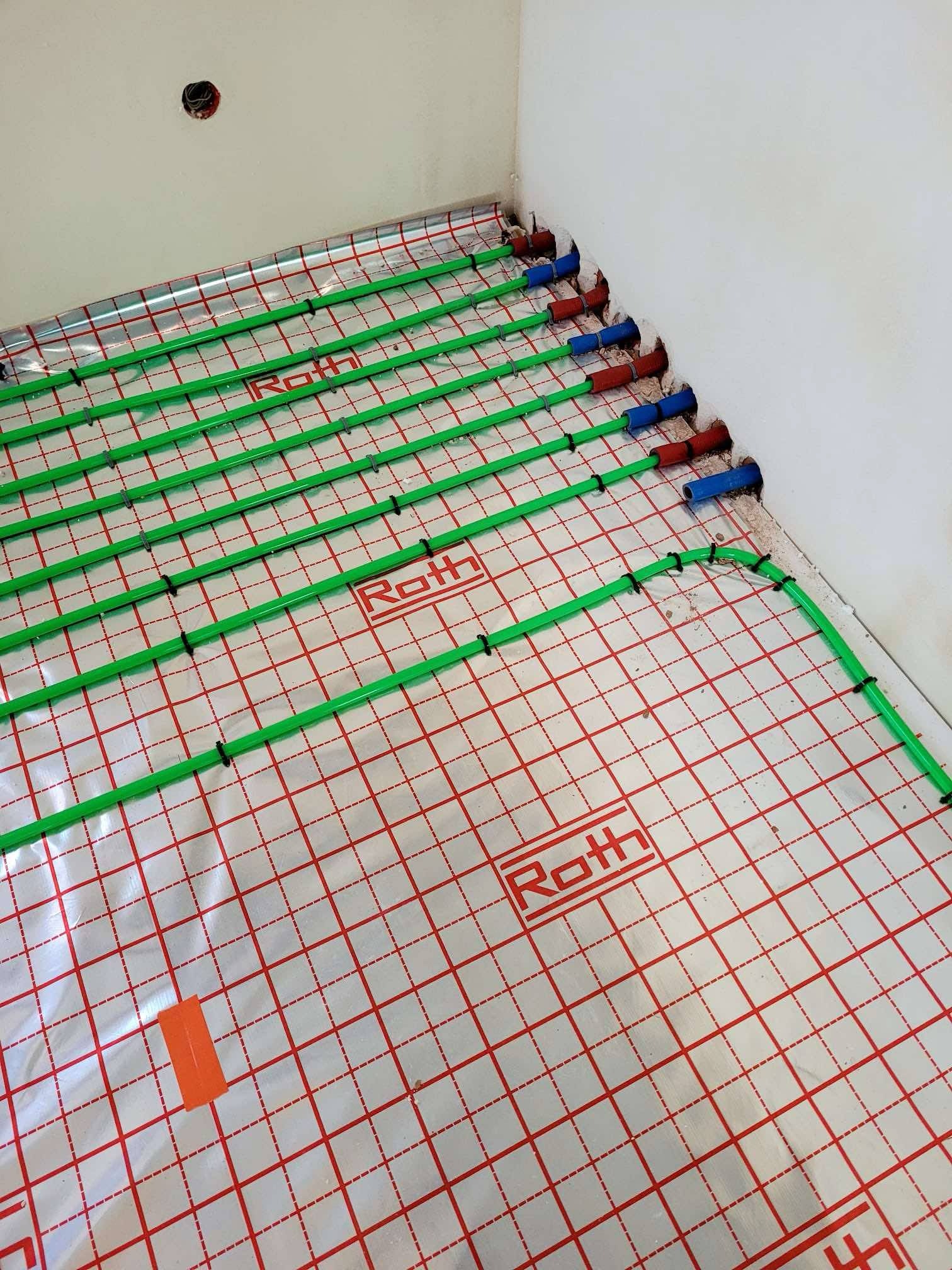 Ułożone zielone rury ogrzewania podłogowego Roth na folii z czerwonym nadrukiem kratki w rogu pomieszczenia. Widoczne niebieskie i czerwone końcówki rur przy ścianie.
