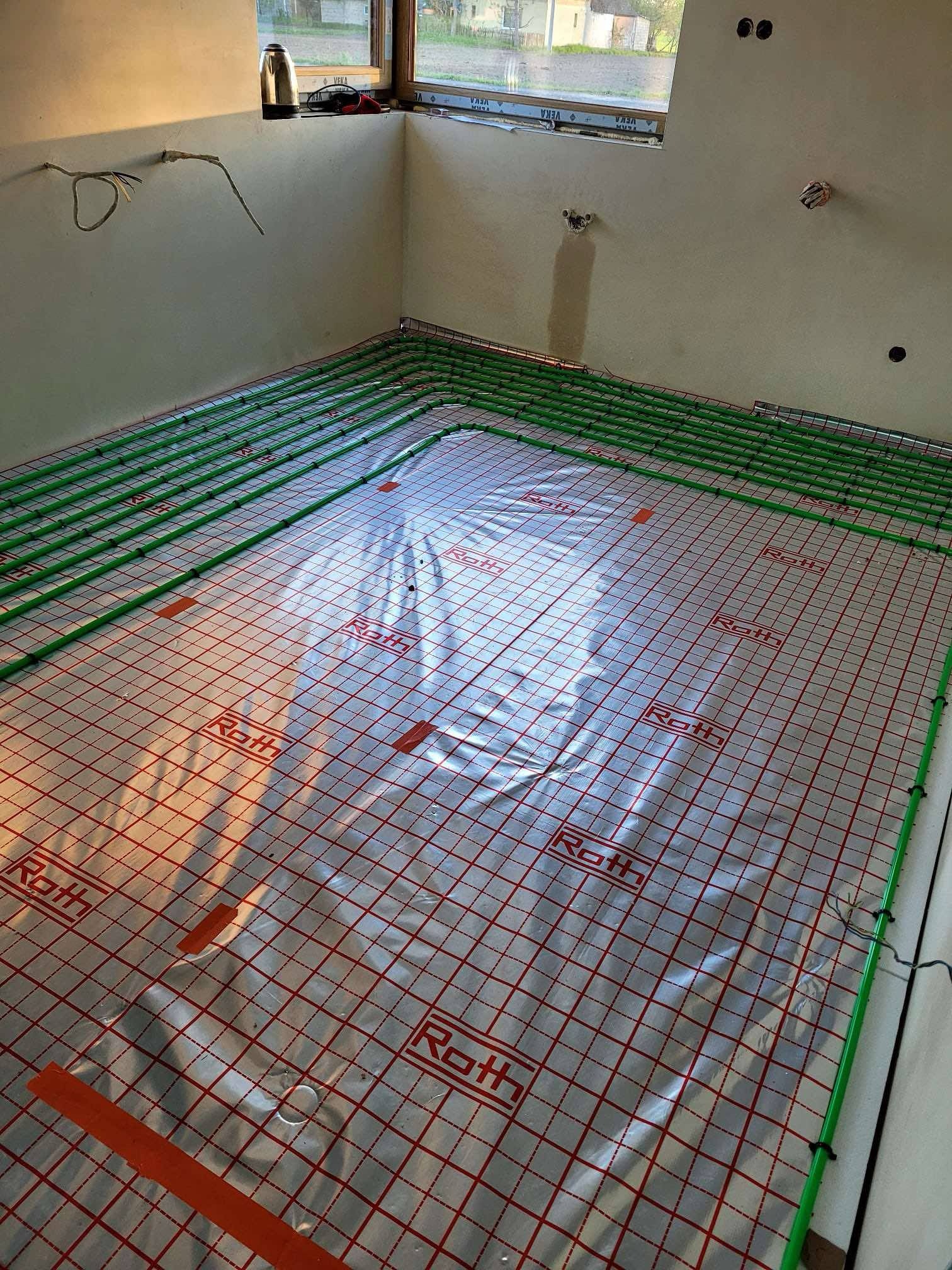 Instalacja ogrzewania podłogowego Roth: zielone rury grzewcze ułożone na folii z czerwoną siatką w surowym wnętrzu. Widoczne elementy instalacji elektrycznej na ścianie.