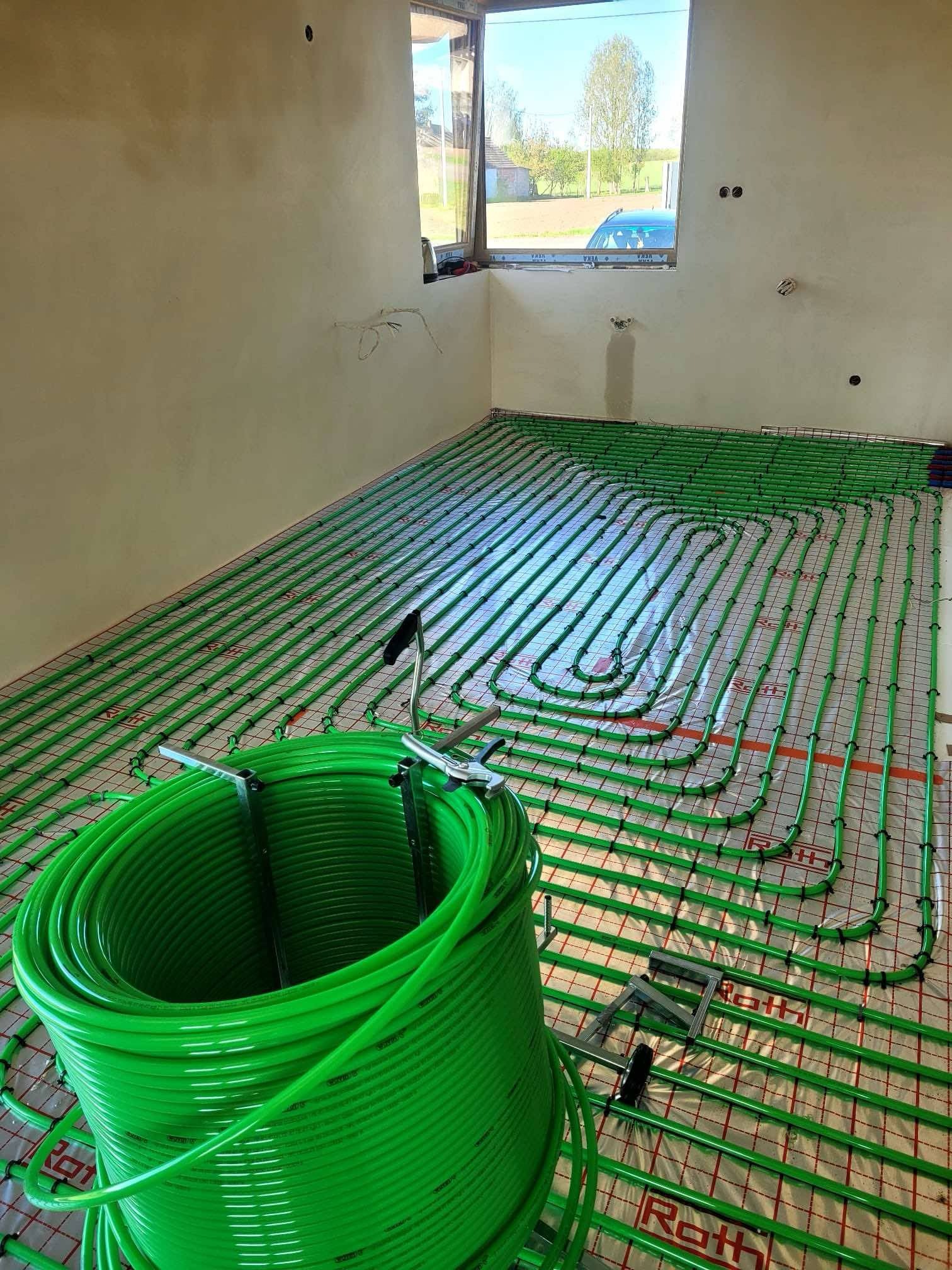Zielone rury ogrzewania podłogowego Roth, ułożone na folii z siatką, w surowym wnętrzu z oknem, przed wylaniem posadzki. Część rury w zwoju widoczna z przodu.