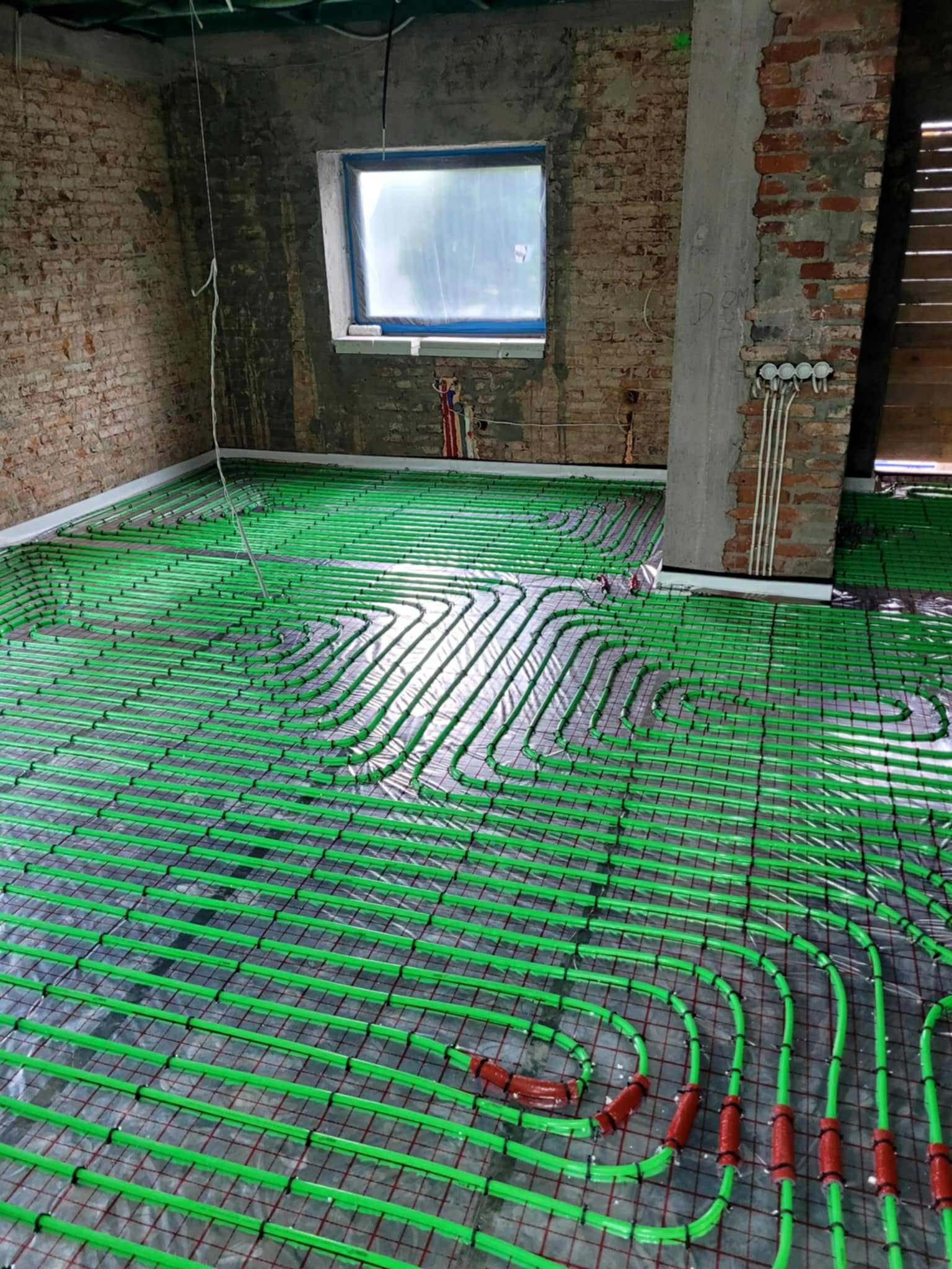 Wnętrze z instalacją ogrzewania podłogowego z zielonych rur na folii, widoczne ściany z czerwonej cegły i okno z folią ochronną. Etap przed wylewką.