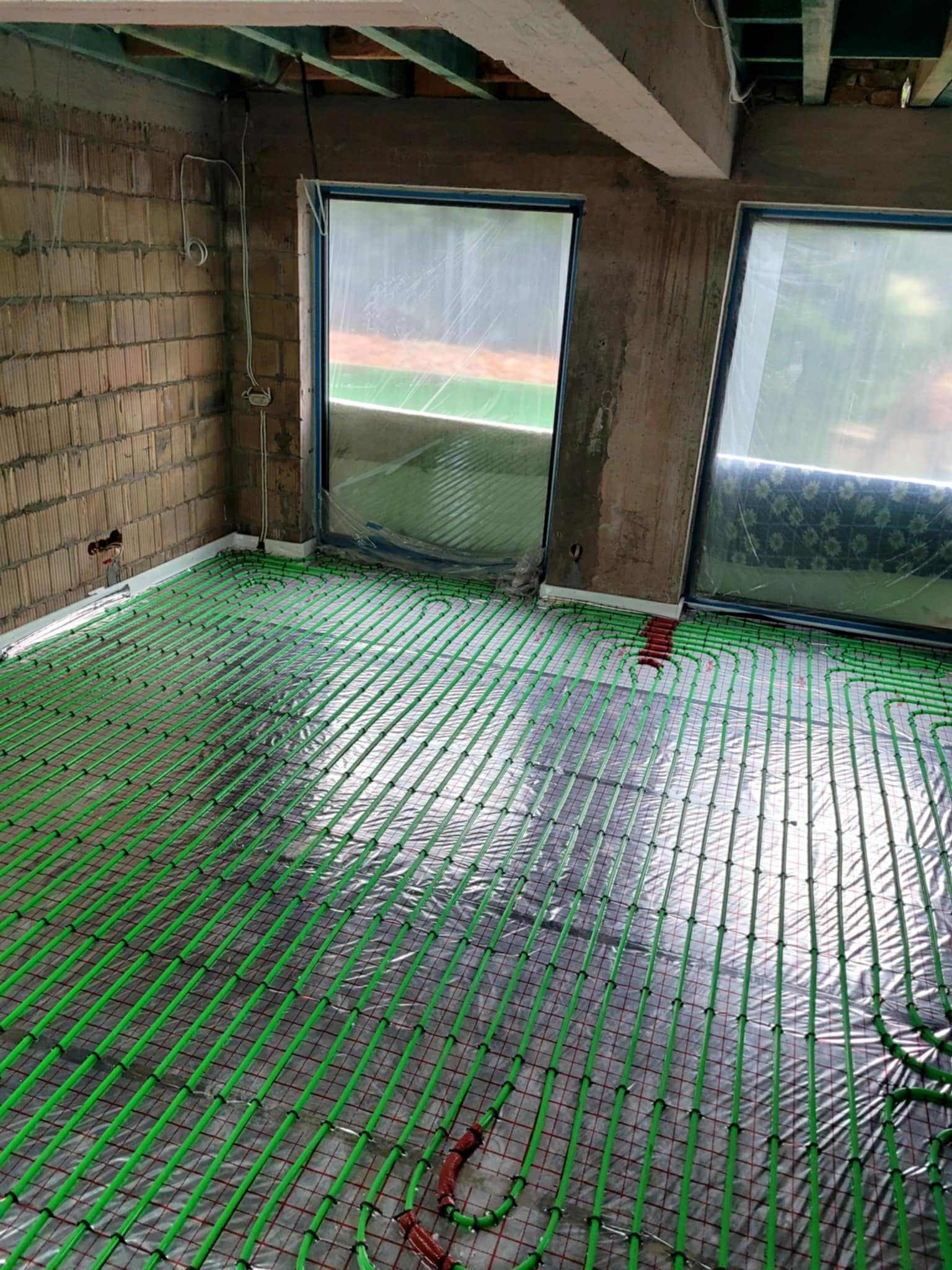 Zielone rury ogrzewania podłogowego ułożone na folii odblaskowej w surowym wnętrzu z oknami, przygotowane do zalania wylewką. Widok z góry.