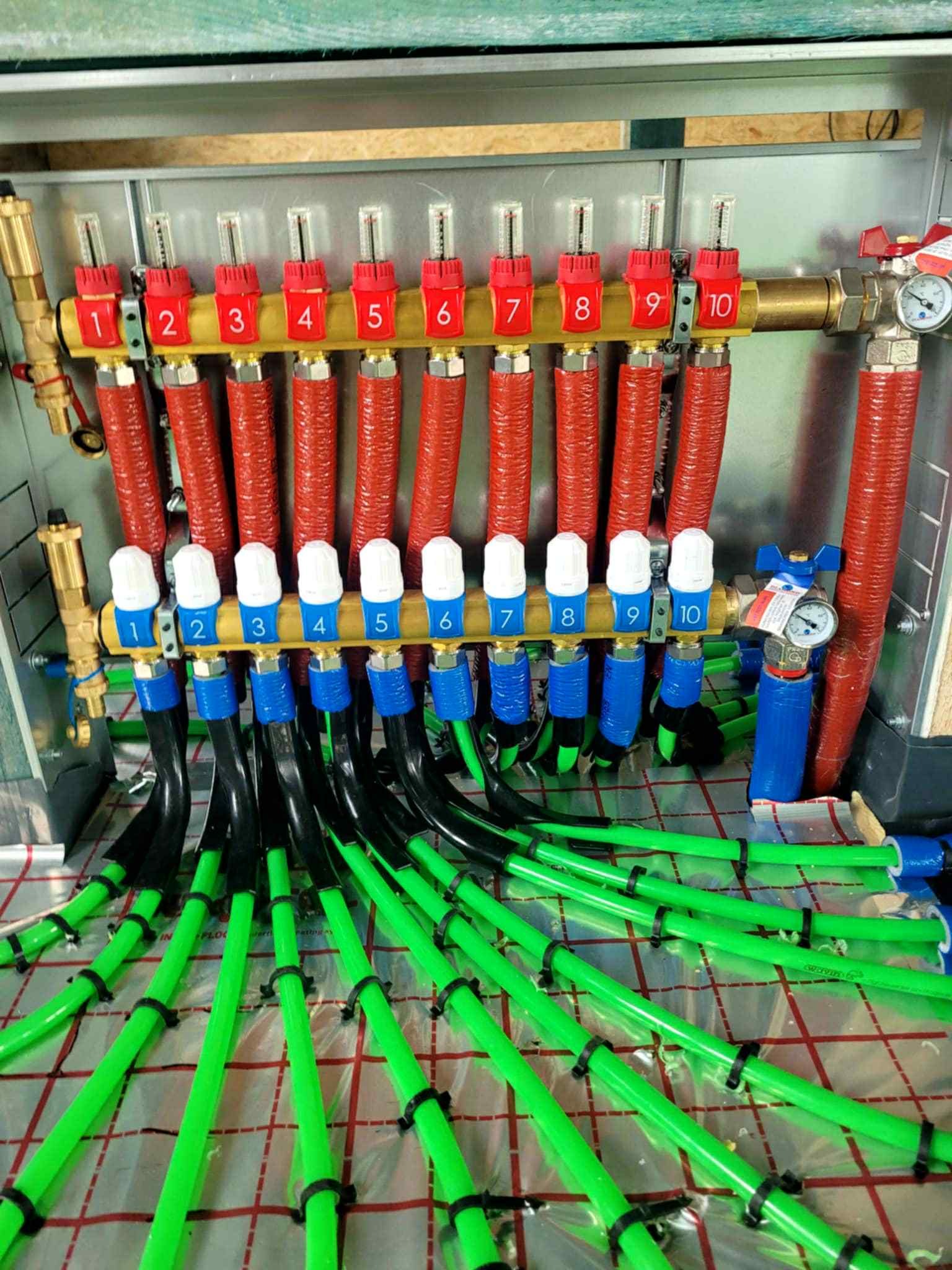 Rozdzielacz ogrzewania podłogowego z dziesięcioma obwodami, z czerwonymi i niebieskimi zaworami oraz zielonymi rurami, zamocowanymi czarnymi opaskami, na folii izolacyjnej.