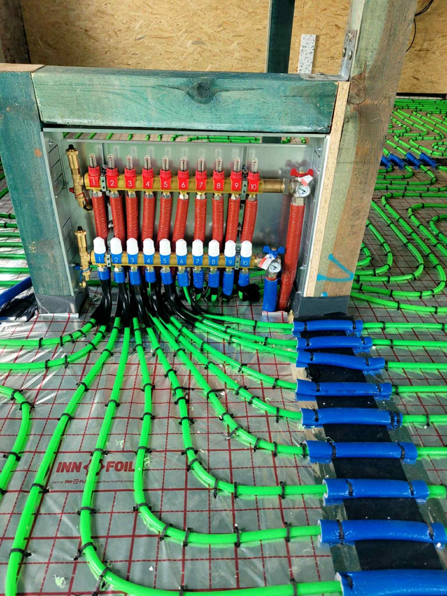 Rozdzielacz ogrzewania podłogowego z zielonymi rurami, przymocowanymi czarnymi opaskami do folii INN FOIL z czerwoną siatką, widok z góry.