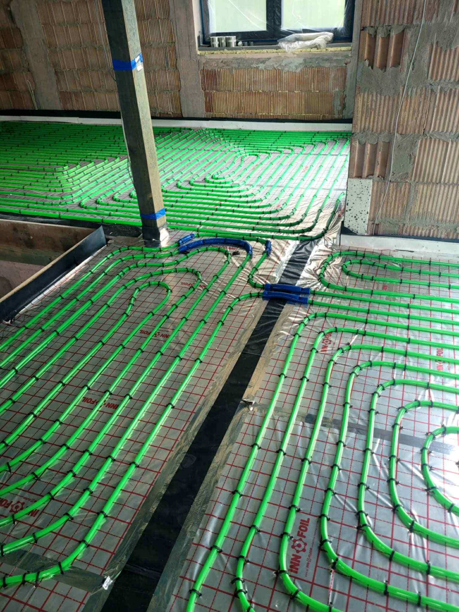 Zielone rury ogrzewania podłogowego ułożone spiralnie na folii z rastrem, widoczne elementy konstrukcji dachu i ściany z pustaków.