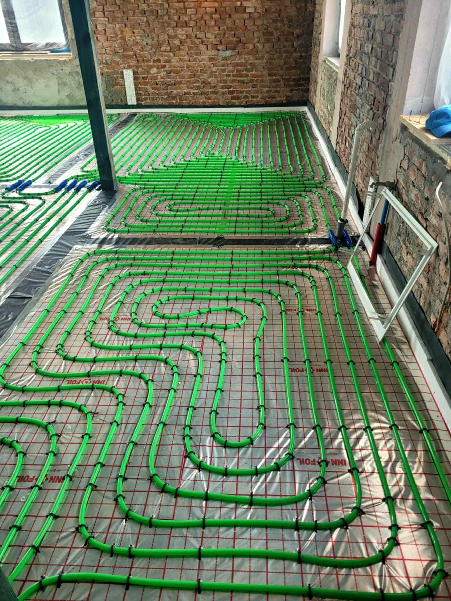 Zielone rury ogrzewania podłogowego ułożone w pętle na folii z siatką, widoczne elementy montażowe i surowa cegła w tle. Instalacja w trakcie realizacji.