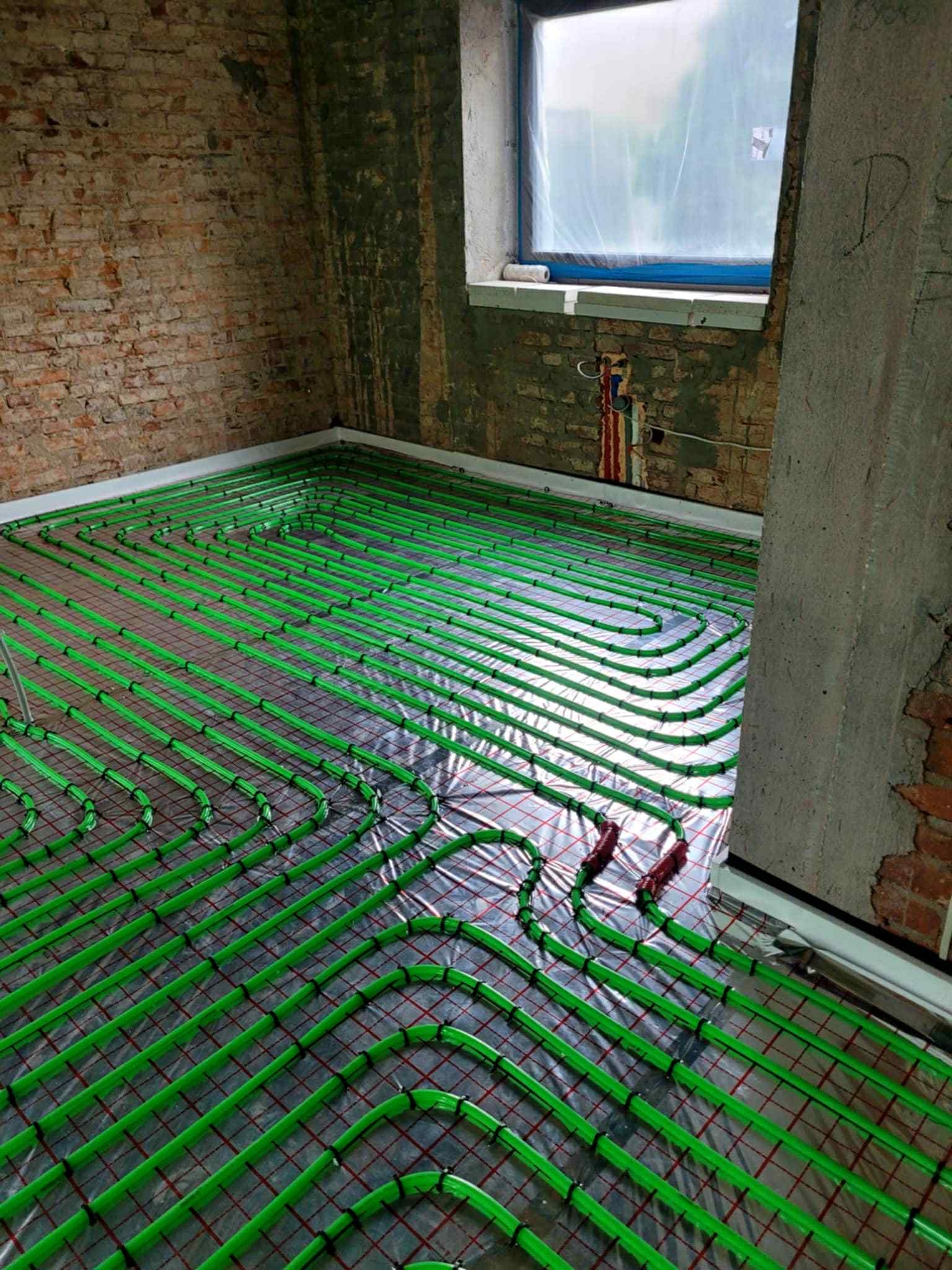 Zielone rury ogrzewania podłogowego ułożone na folii w surowym wnętrzu z ceglaną ścianą i oknem zabezpieczonym folią malarską. Etap instalacji przed wylewką.