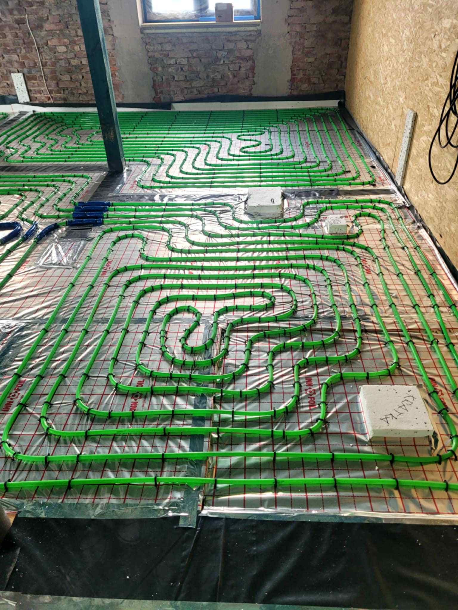 Zielone rury ogrzewania podłogowego ułożone na folii z siatką w surowym wnętrzu z ceglaną ścianą i drewnianą boazerią. Widok z góry na instalację przed wylewką.