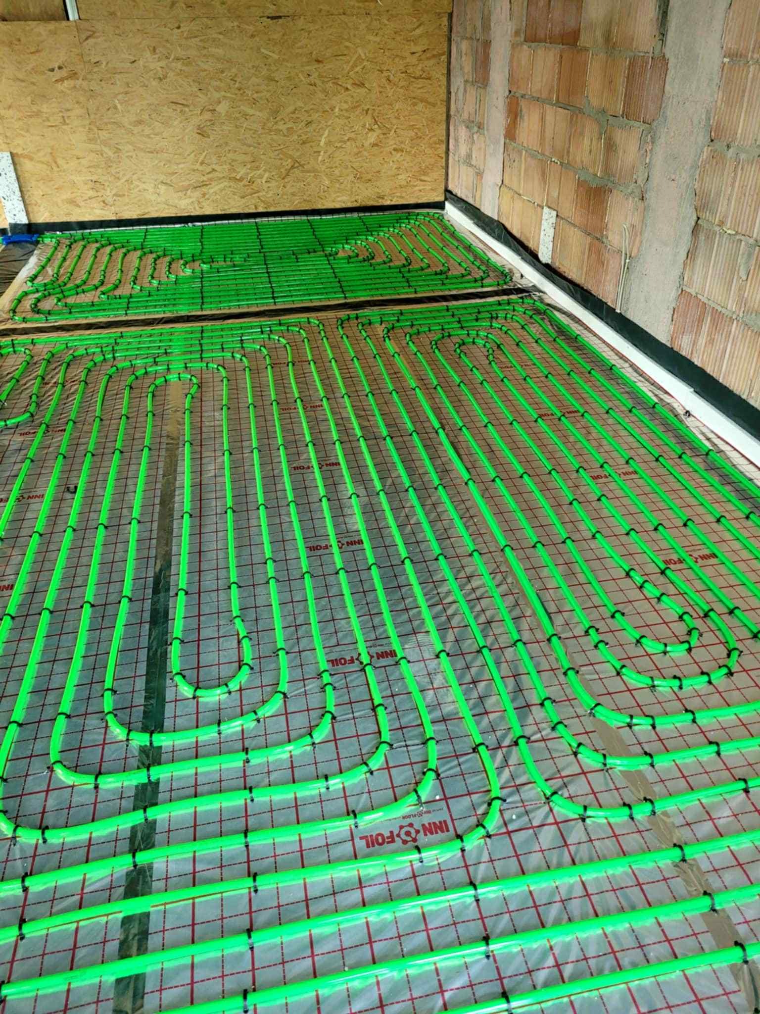 Układanie zielonych rur ogrzewania podłogowego na folii INN-FOIL, widok z góry na tle surowych ścian i płyty OSB. Elementy instalacji przymocowane czarnymi uchwytami.