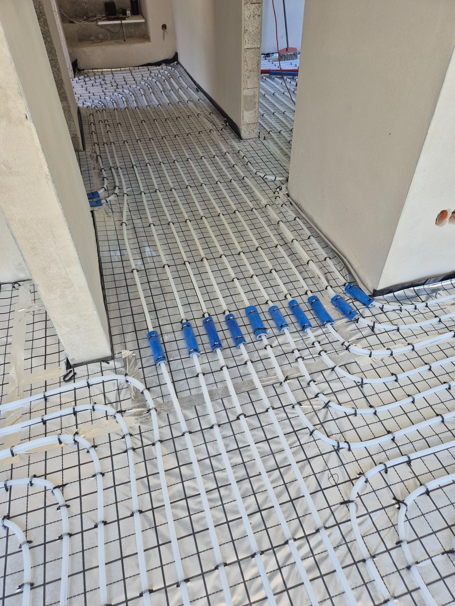 Białe rury ogrzewania podłogowego, mocowane do czarnej siatki na srebrnej folii izolacyjnej, widoczne niebieskie elementy łączeniowe, surowy stan wykończenia.