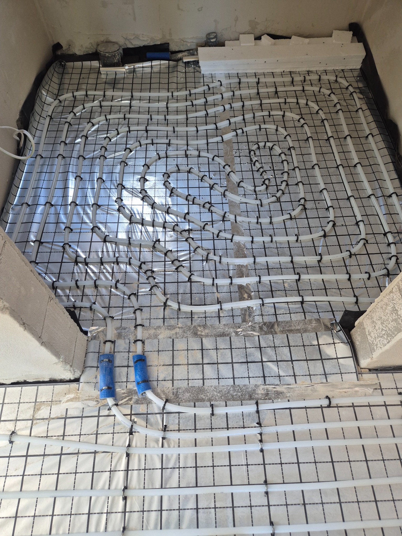 Białe rury ogrzewania podłogowego ułożone spiralnie na folii z siatką, zabezpieczone czarnymi opaskami. Fragment ściany i niebieskie osłony na rurach.