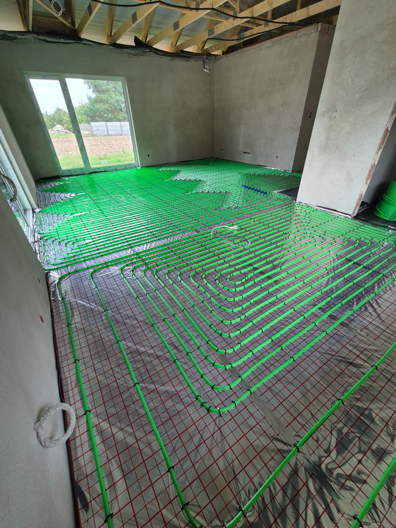 Zielone rury ogrzewania podłogowego ułożone na folii z czerwoną kratką w surowym wnętrzu z widoczną konstrukcją dachu i oknem z widokiem na zewnątrz.