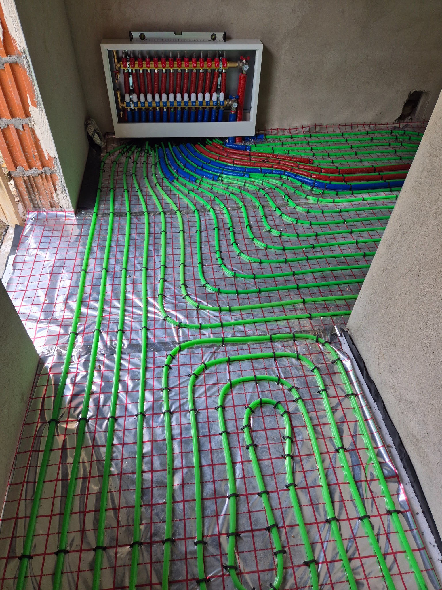 Rozłożone rury ogrzewania podłogowego w kolorze zielonym, niebieskim i czerwonym na folii z siatką, widoczna szafka rozdzielcza z zaworami i poziomica.