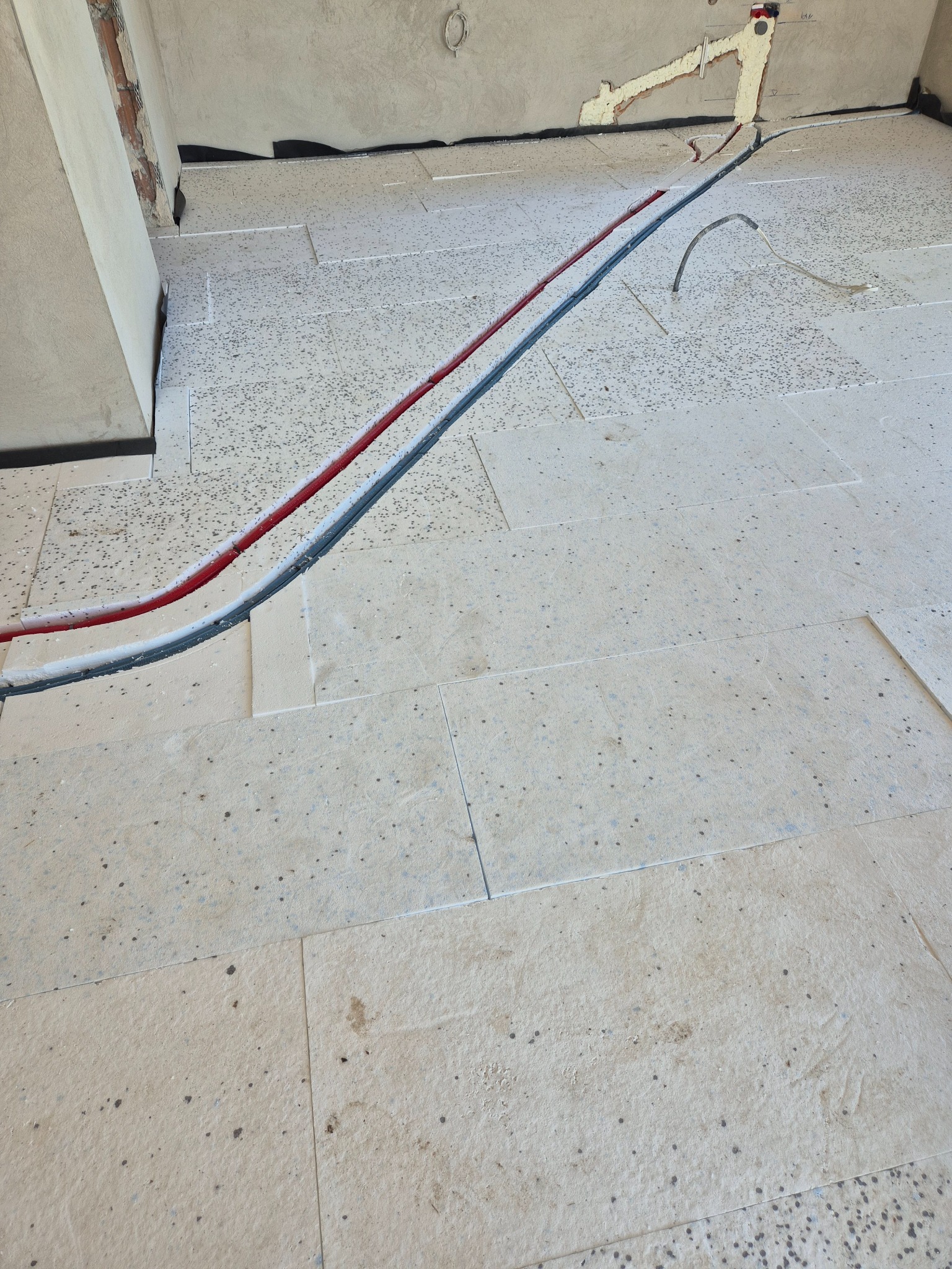 Przygotowanie do ogrzewania podłogowego: izolacja z płyt styropianowych z wyfrezowanymi kanałami na rury grzewcze, częściowo ułożone przewody w kolorze czerwonym i niebieskim.