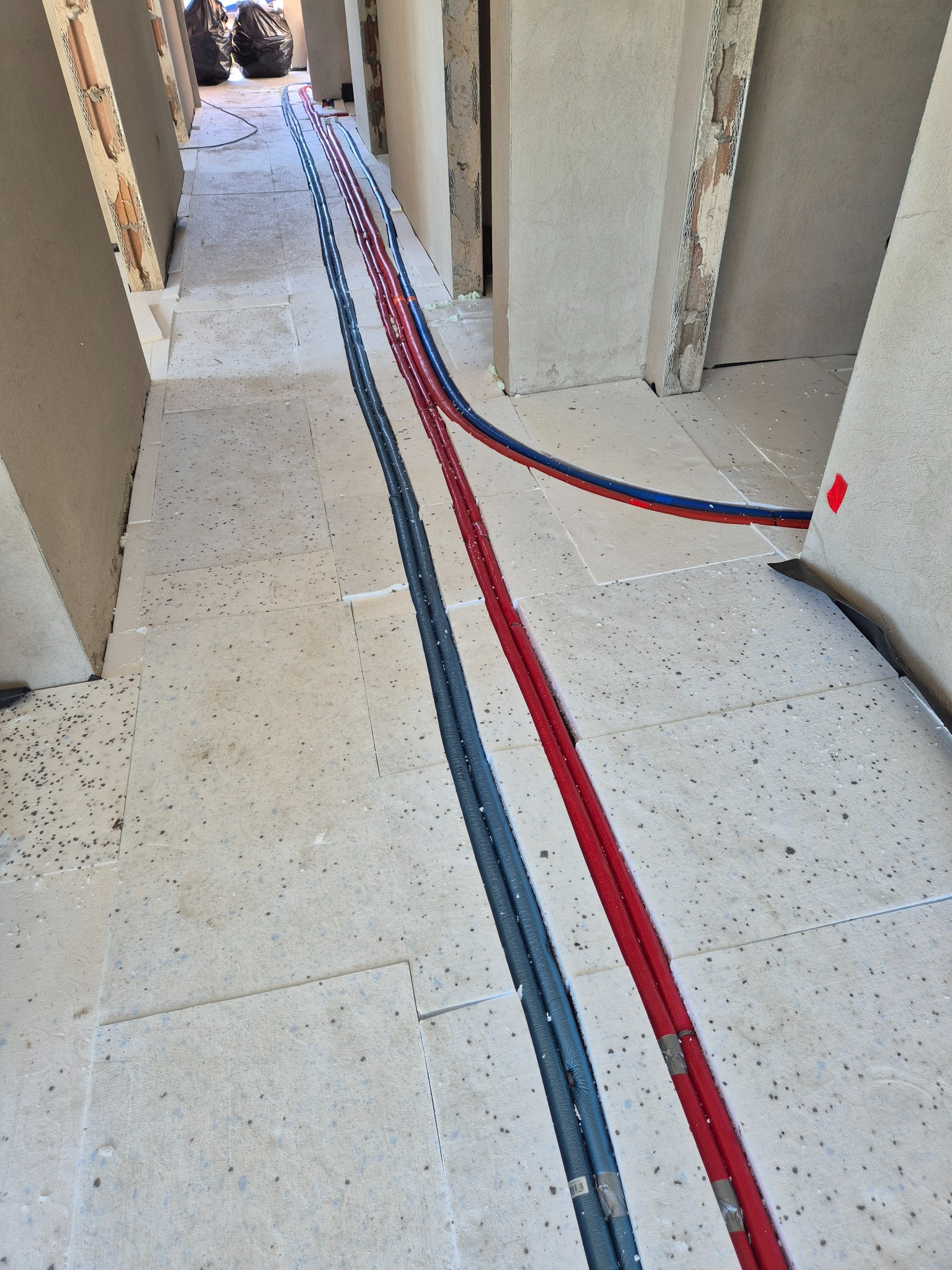 Instalacja rur grzewczych (czerwone i niebieskie) oraz wodnych (szare) na izolacji podłogowej w korytarzu podczas remontu. Widoczne przygotowania do wylewki.