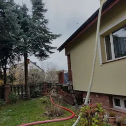 Proces ocieplania domu z użyciem węża doprowadzającego materiał izolacyjny do poddasza, widoczny wąż na elewacji i na trawniku w ogrodzie