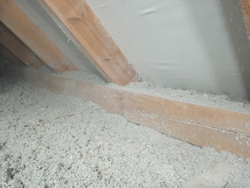 Poddasze ocieplone szarą wełną celulozową, widoczne drewniane krokwie i folia paroizolacyjna, oświetlenie naturalne.