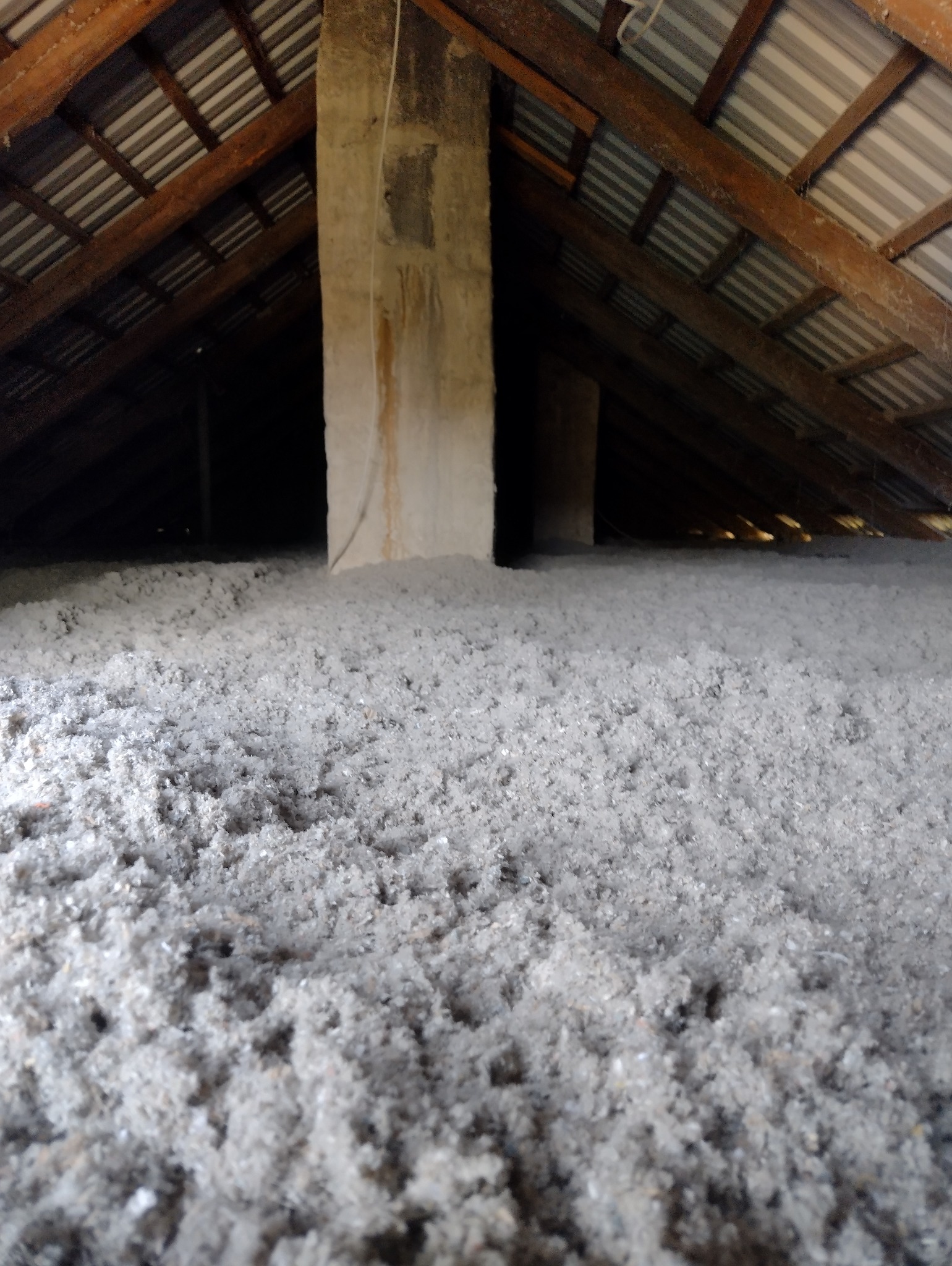 Poddasze z widoczną drewnianą konstrukcją dachu i kominem, wypełnione szarą, sypką izolacją.