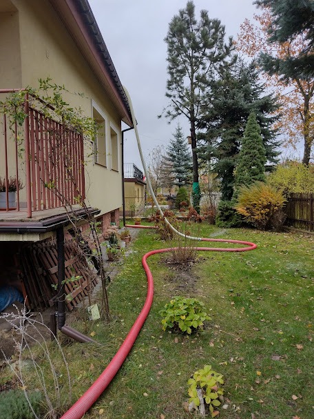 Czerwony wąż doprowadzający materiał do ocieplania poddasza, biegnący przez trawnik obok budynku z balkonem, widok na elewację i drzewa w tle.