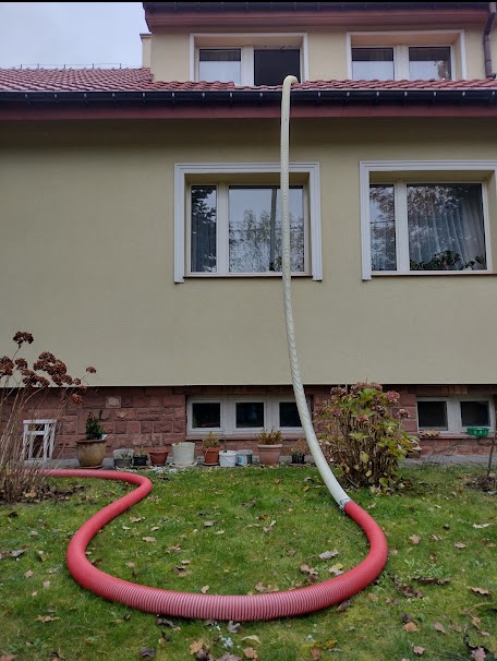 Węże do wdmuchiwania izolacji prowadzone z zewnątrz budynku przez okno poddasza, zielony trawnik w jesiennych liściach.