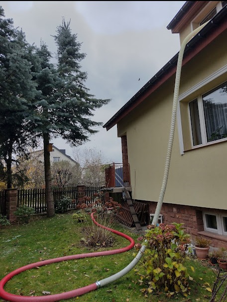 Proces ocieplania domu z użyciem węża doprowadzającego materiał izolacyjny do poddasza, widoczny wąż na elewacji i na trawniku w ogrodzie