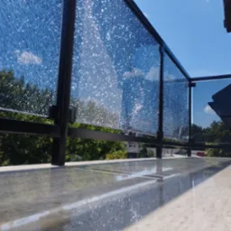 Balkon z nowoczesną balustradą z czarnych profili i szkła ornamentowego, odbijającego niebo i zieleń drzew w słoneczny dzień.