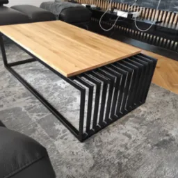 Stolik kawowy z blatem z jasnego drewna i czarną, metalową podstawą o geometrycznym designie, na tle szarego dywanu i czarnej sofy.