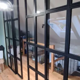 Przeszklona ścianka działowa z czarnymi metalowymi profilami, oddzielająca przestrzeń na dwóch poziomach, widok na schody i okno dachowe.