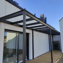 Nowoczesna pergola z czarnej stali z przezroczystym zadaszeniem, przylegająca do białego budynku, widoczne butle z gazem w tle.