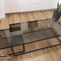 Stolik kawowy z czarną metalową ramą i szklanym blatem, ustawiony na drewnianej podłodze, obok doniczki z rośliną. Widoczne białe szafki w tle.