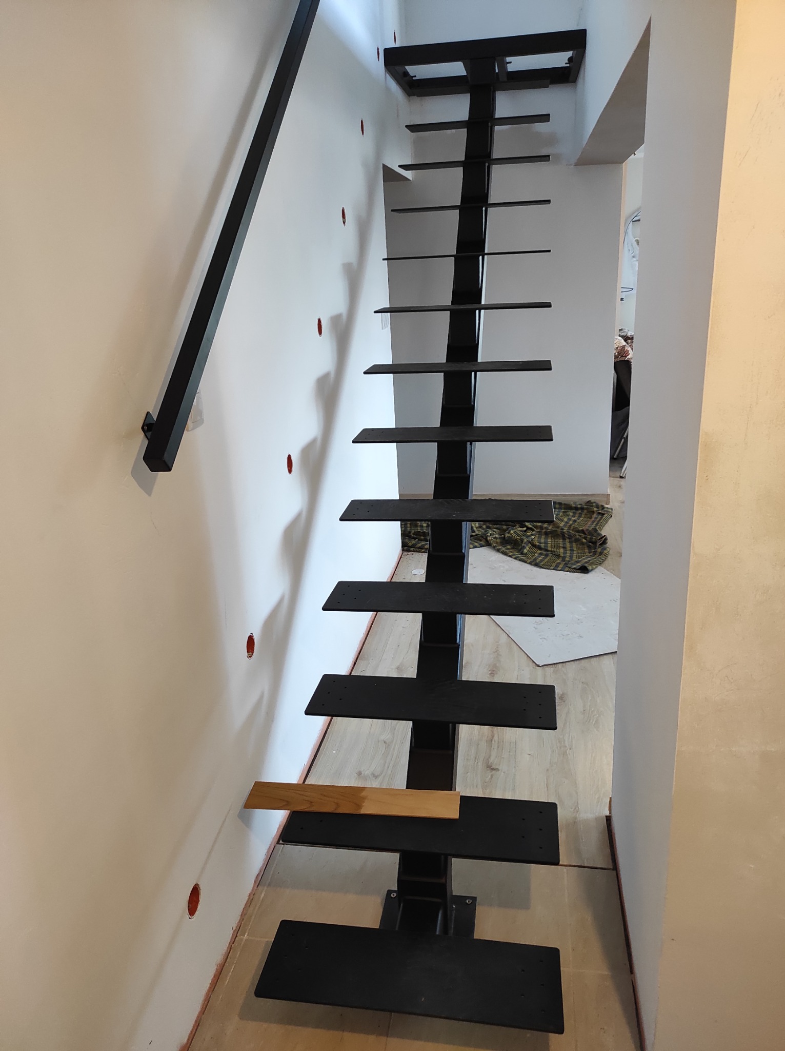 Metalowe schody ażurowe w trakcie montażu, z czarną konstrukcją i jednym drewnianym stopniem, widoczne ślady prac wykończeniowych na ścianie i podłodze.