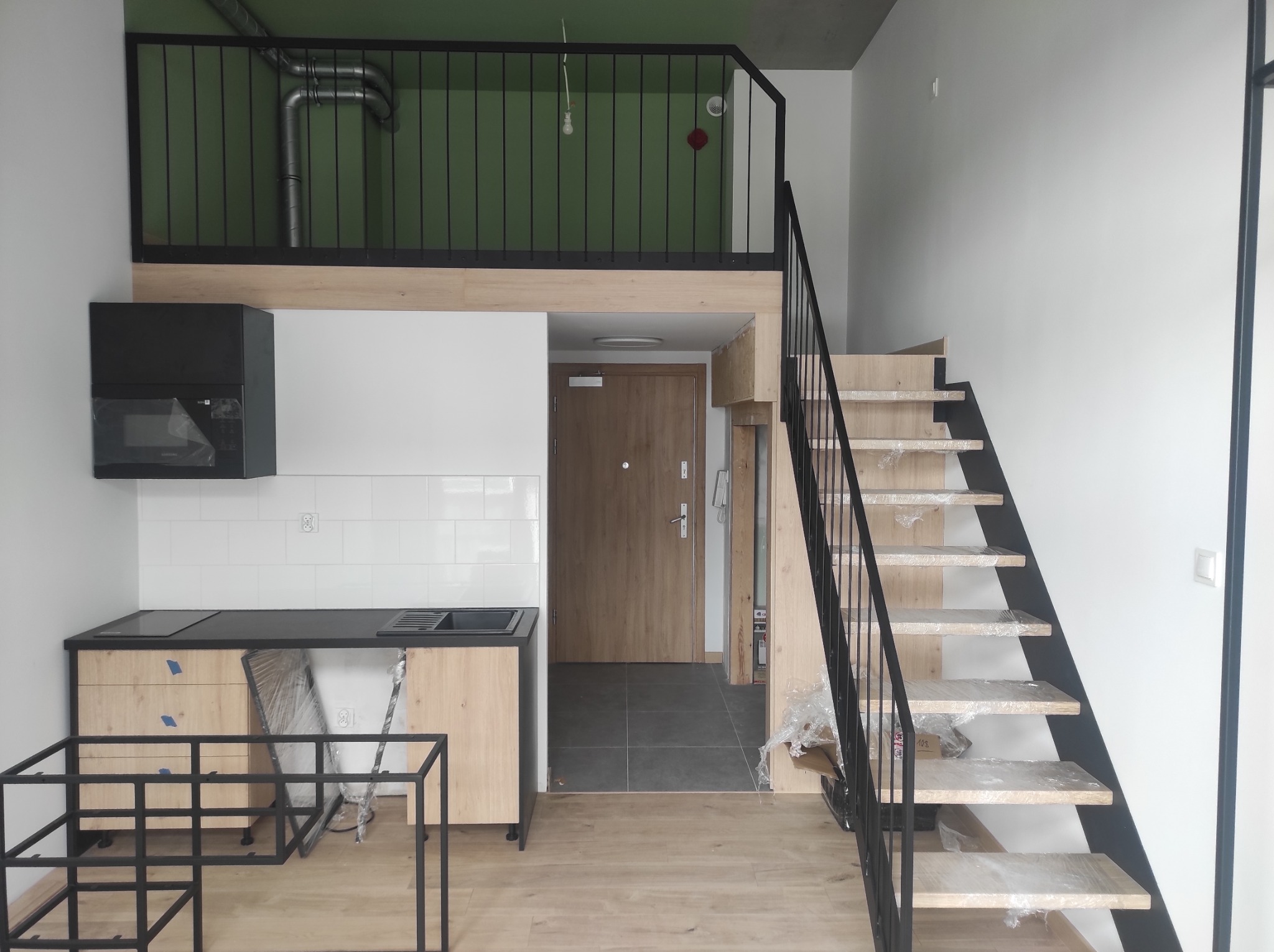 Wnętrze mieszkania z antresolą, drewniane schody z czarną metalową balustradą, kuchnia z zabudową w trakcie montażu, widoczne elementy konstrukcji metalowej.