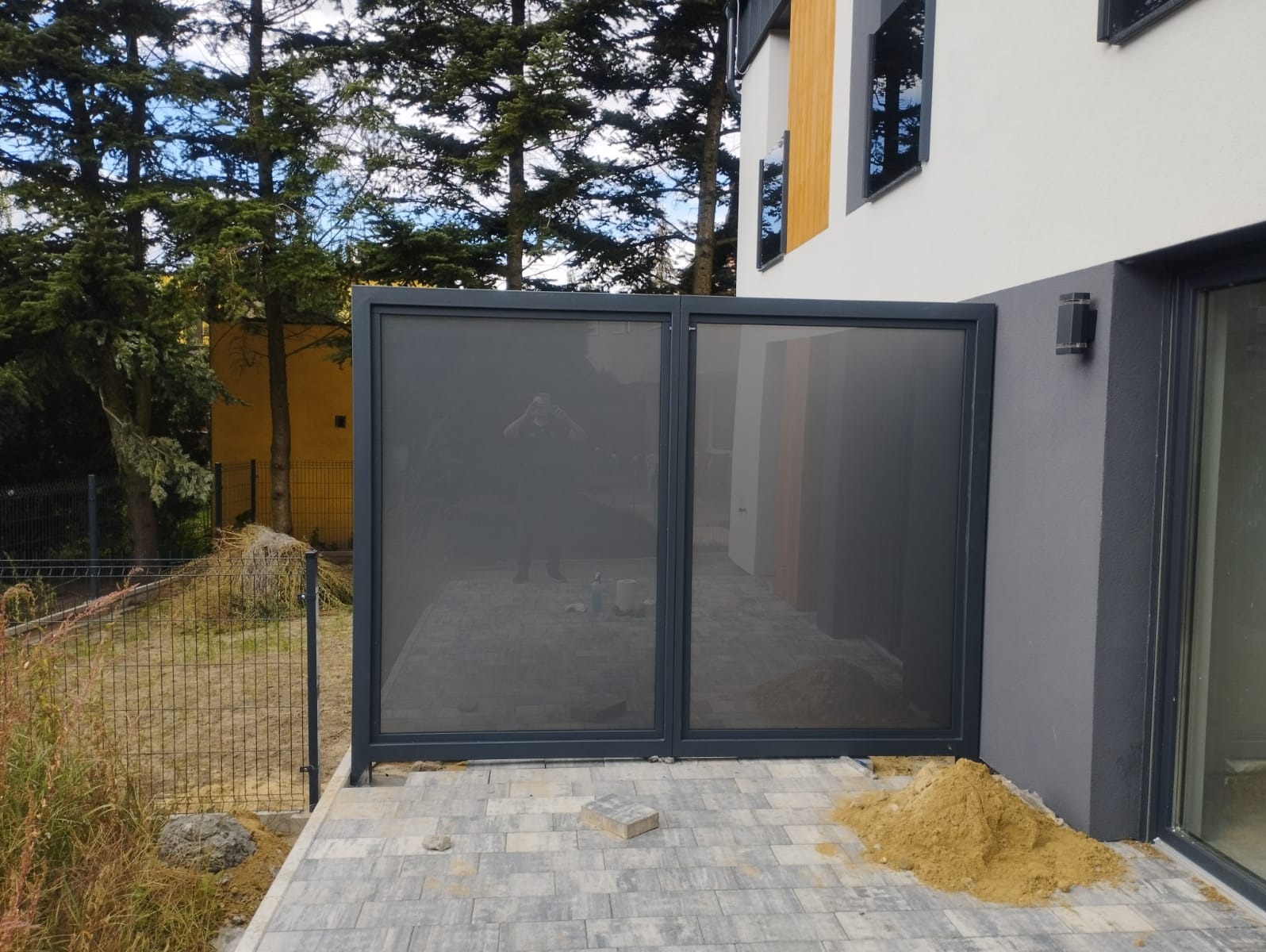 Nowoczesna, dwuczęściowa przesłona tarasowa z ciemną ramą, zapewniająca prywatność, z widocznym odbiciem fotografa, ustawiona na szarej kostce brukowej, z usypaną kupką piasku obok