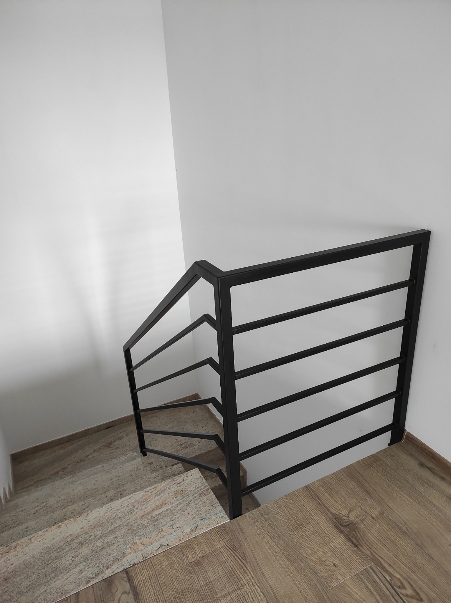 Minimalistyczna, czarna poręcz schodowa z poziomymi prętami, widok z góry na schody wyłożone płytkami imitującymi kamień i podłogę z paneli drewnopodobnych, białe ściany.