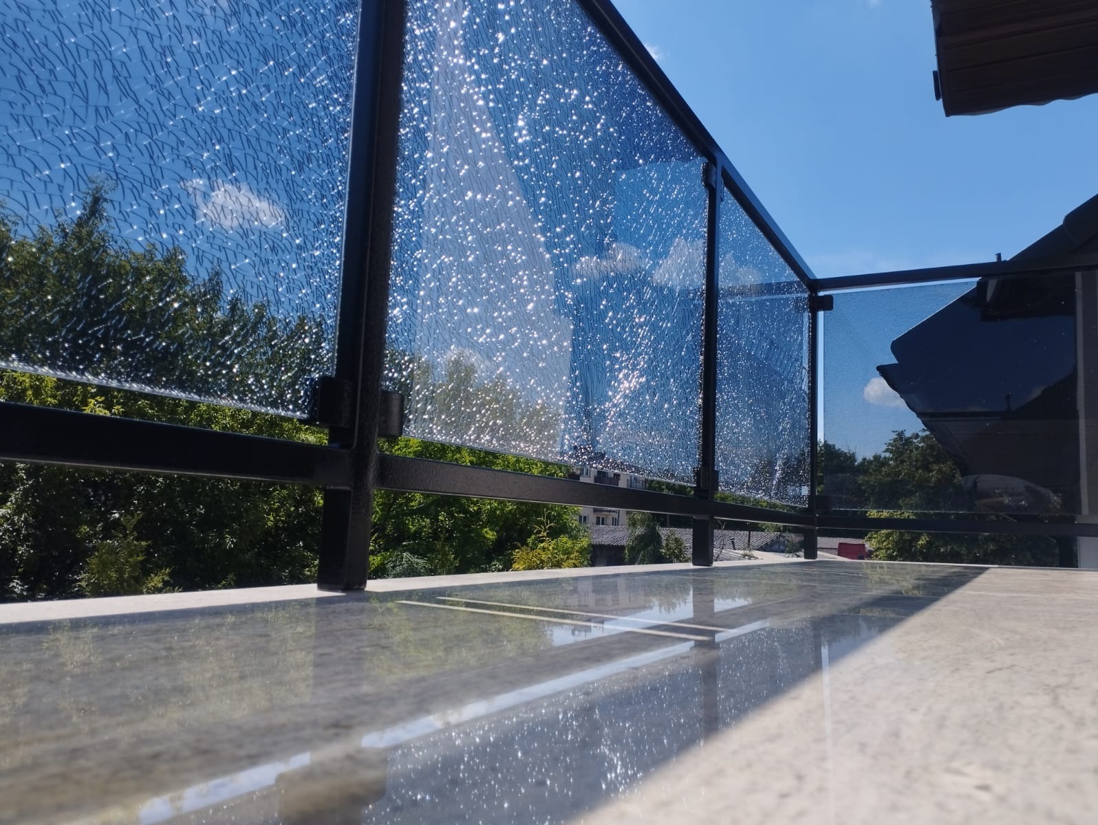 Balkon z nowoczesną balustradą z czarnych profili i szkła ornamentowego, odbijającego niebo i zieleń drzew w słoneczny dzień.