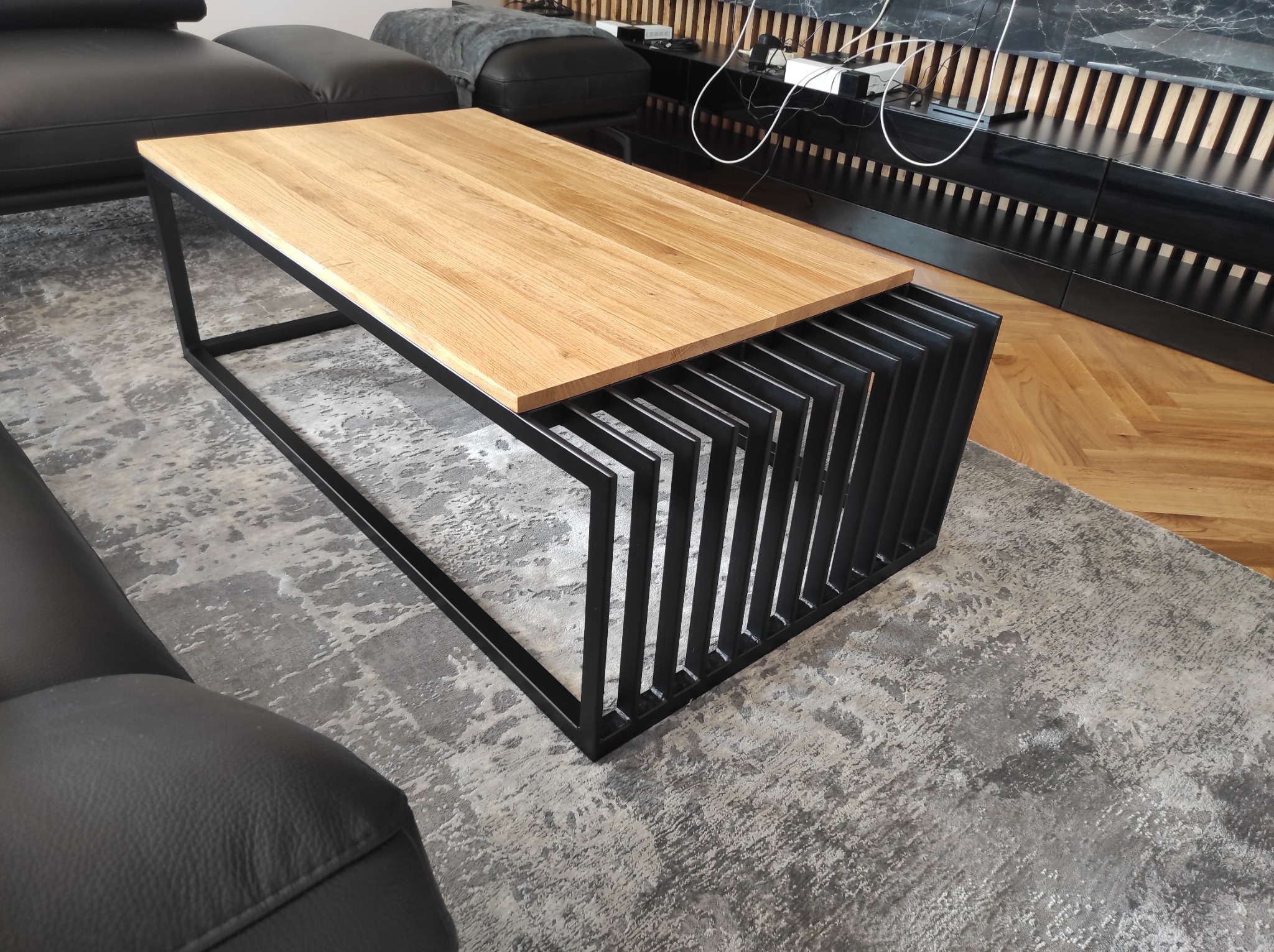 Stolik kawowy z blatem z jasnego drewna i czarną, metalową podstawą o geometrycznym designie, na tle szarego dywanu i czarnej sofy.